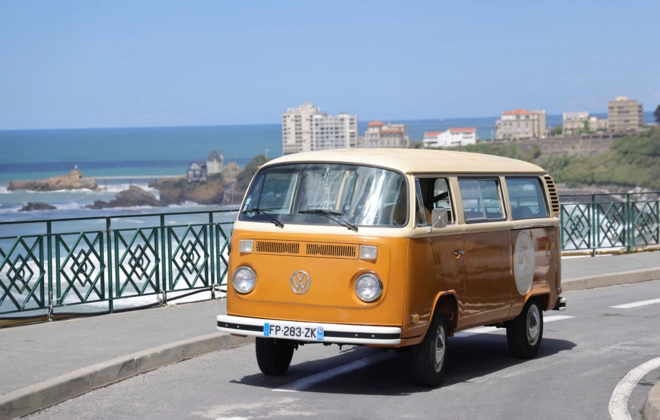 Volkswagen Combi vintage orange et beige circulant sur la corniche de Biarritz, avec en arrière-plan la Villa Belza sur son rocher, le front de mer et l'océan Atlantique sous un ciel bleu ensoleillé.