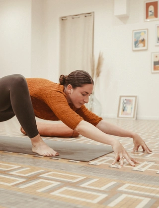 Femme en pull terracotta et legging noir effectuant une posture de yoga en fente basse sur un tapis, dans un studio lumineux décoré de cadres et d'herbes de la pampa.