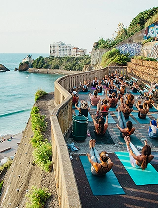 Cours de yoga en plein air sur une terrasse en bord de falaise à Biarritz, avec un grand groupe de participants sur tapis colorés face à l'océan Atlantique et la Villa Belza en arrière-plan.