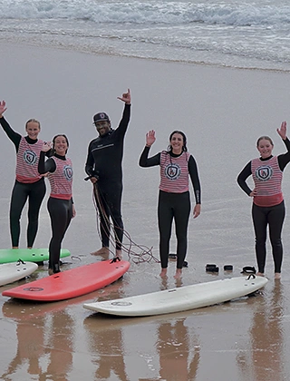 Groupe de quatre élèves en combinaison de surf et chasuble rayée posant avec leur moniteur sur la plage, les bras levés, devant leurs planches colorées au bord de l'eau.
