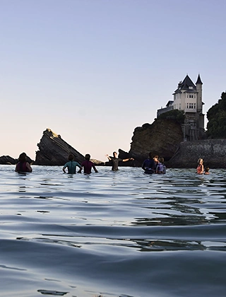 Baigneurs dans l'océan au crépuscule face à la Villa Belza de Biarritz, manoir néogothique perché sur son rocher, sous un ciel bleu pâle aux tons rosés.