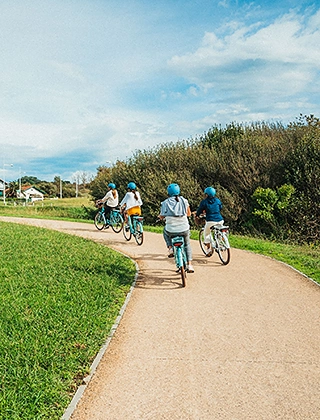 Groupe de quatre cyclistes portant des casques bleus vus de dos sur une piste cyclable longeant un espace vert, sous un ciel nuageux en bord de côte basque.