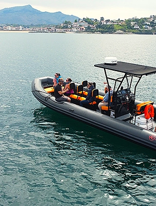 Groupe de passagers à bord d'un semi-rigide gris aux coussins orange naviguant sur une baie calme, avec en arrière-plan le littoral et les montagnes basques.