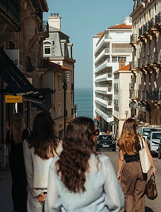 Trois femmes vues de dos se promenant dans une rue commerçante de Biarritz bordée d'immeubles haussmanniens, avec une échappée sur l'océan Atlantique en fond de perspective.