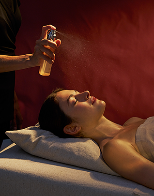 Femme allongée les yeux fermés sur une table de soin, un thérapeute vaporise un brume parfumée au-dessus de son visage, dans une atmosphère enveloppante aux tons bordeaux profonds et lumière dorée
