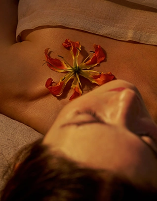 Femme allongée les yeux fermés sur une table de soin, une fleur exotique aux pétales orangés et rouges posée sur son dos, dans une atmosphère sereine et sensorielle aux tons chauds et terreux