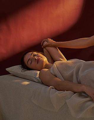 Femme allongée sur le dos les yeux fermés, drapée dans un linge blanc, le bras étiré tenu par les mains d'un thérapeute, dans une atmosphère enveloppante aux tons bordeaux et lumière dorée rasante