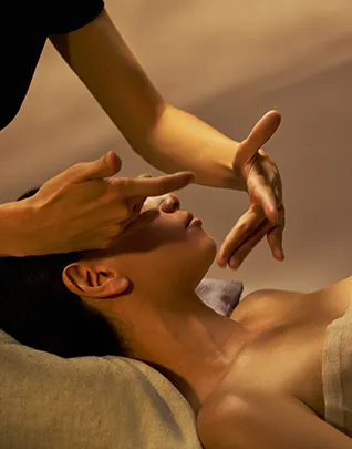 Personne allongée les yeux fermés recevant un massage du visage, les deux mains du thérapeute exerçant des pressions symétriques sur le front et les tempes, dans une lumière dorée et tamisée aux tons ambrés