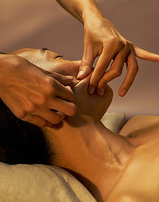 Gros plan sur un massage du visage et de la mâchoire, les mains du thérapeute travaillant avec précision sur le cou et le menton d'une personne allongée, dans une lumière dorée et chaude aux tons ocre