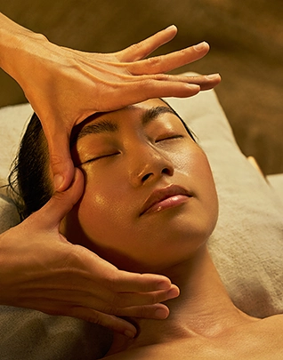 Femme aux yeux fermés recevant un massage du visage, une main glissant sur le front et l'autre soutenant la nuque, dans une atmosphère intimiste aux tons dorés et bruns profonds