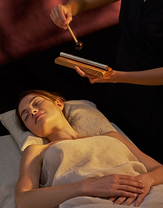 Femme allongée les yeux fermés sur une table de soin, un thérapeute tient au-dessus d'elle un diapason qu'il s'apprête à frapper avec un maillet, dans une ambiance sombre et intimiste aux reflets dorés et bordeaux