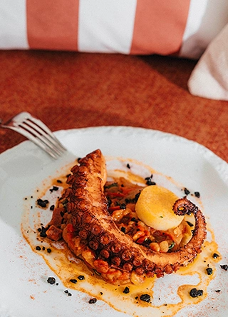 Tentacule de pieuvre grillée dressée en spirale sur une sauce safranée, accompagnée d'une quenelle de crème citronnée et de légumes confits, servie sur assiette blanche.