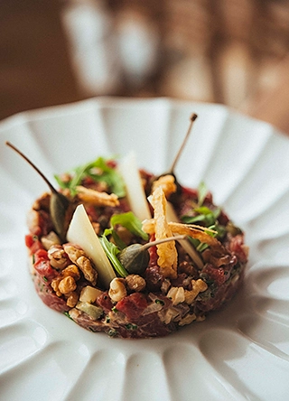 Tartare de bœuf dressé en cercle, garni de noisettes, câpres, copeaux de parmesan et chips croustillantes, sur assiette blanche striée