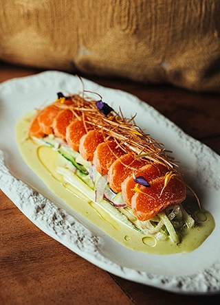 Pavé de saumon mi-cuit en tranches sur lit de fenouil et sauce verte, agrémenté de paille croustillante et de fleurs violettes, servi sur assiette oblongue blanche.