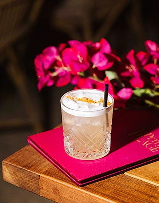 Cocktail glacé servi dans un verre taillé avec une paille noire et saupoudré d'épices dorées, posé sur une carte de menu rose fuchsia sur une table en bois, devant un bouquet de fleurs roses fuchsia en arrière-plan