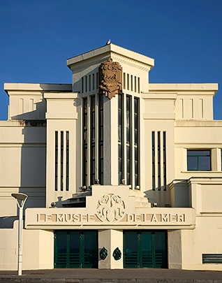 Façade Art déco du Musée de la Mer, bâtiment blanc aux lignes géométriques avec un blason sculpté en son fronton et des portes vertes, photographié sous un ciel bleu intense