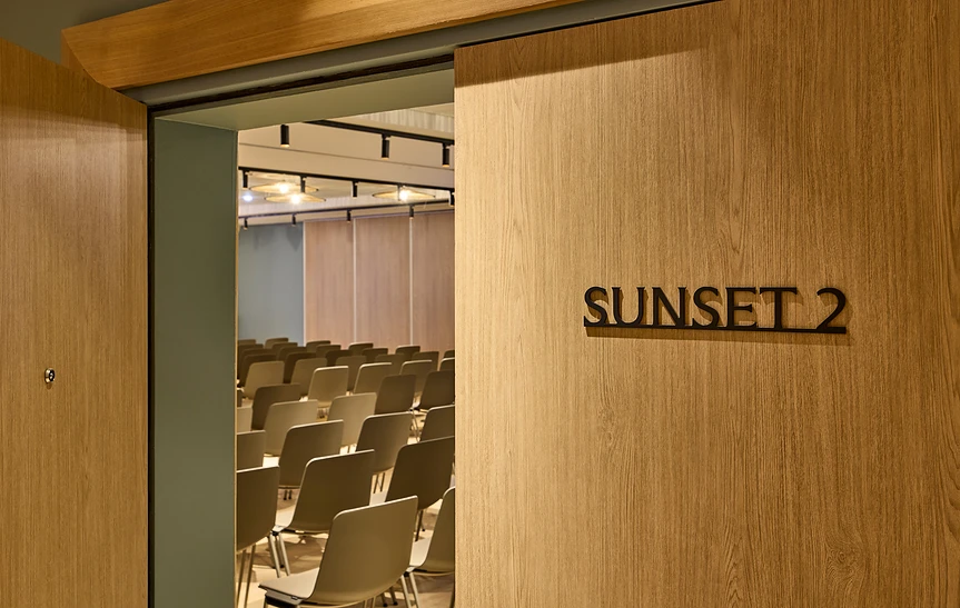 Porte coulissante en bois clair de la salle Sunset 2 de l'hôtel Le Talaia Mgallery Biarritz, entrouverte laissant voir des rangées de chaises grises en configuration théâtre et des cloisons acoustiques en fond de salle