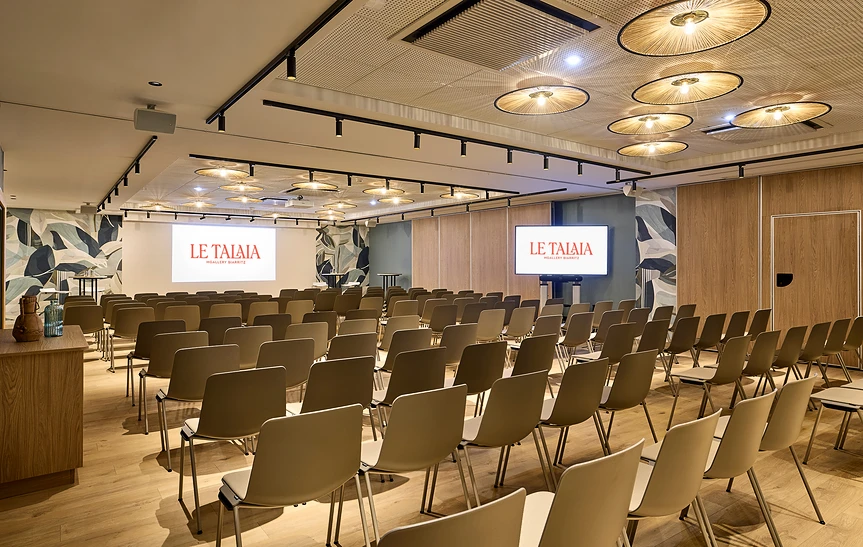 Grande salle de conférence de l'hôtel Le Talaia Mgallery Biarritz en configuration théâtre, vue en angle, avec de nombreuses rangées de chaises grises, deux écrans de projection encadrés de fresques murales végétales, plafond acoustique orné de luminaires ronds en rotin, sol en parquet clair et pupitre en bois sur la gauche