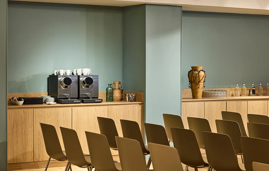 Espace de pause de l'hôtel Le Talaia Mgallery Biarritz, avec buffet en bois clair intégré le long d'un mur vert sauge, équipé de deux machines à café Nespresso, tasses, vases décoratifs et bouteilles d'eau, devant des rangées de chaises grises