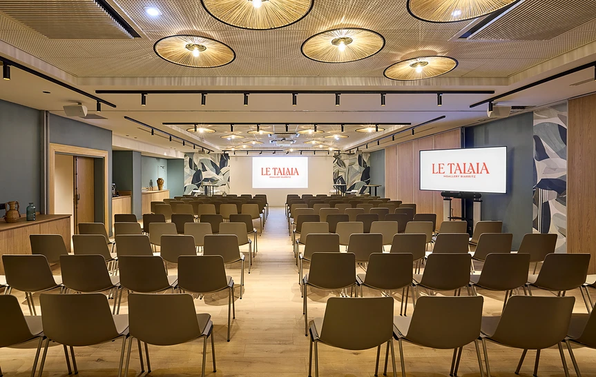 Grande salle de conférence de l'hôtel Le Talaia Mgallery Biarritz en configuration théâtre, vue en perspective centrale, avec rangées de chaises grises disposées en trois allées, un écran de projection au fond et un écran latéral à droite sur pied, fresques murales végétales, luminaires ronds en rotin au plafond, sol en parquet clair et buffet en bois sur la gauche