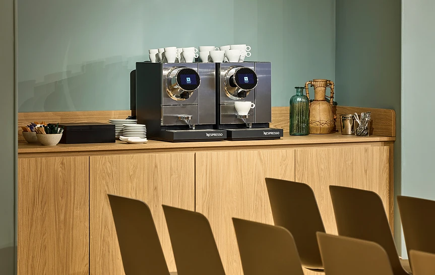 Gros plan sur deux machines à café Nespresso professionnelles posées sur un buffet en bois clair, avec tasses, soucoupes et vases décoratifs, dans l'espace de pause de l'hôtel Le Talaia Mgallery Biarritz