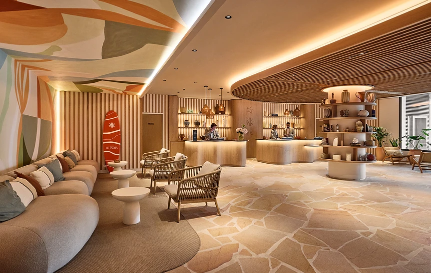 Hall d'accueil de l'hôtel Le Talaia Mgallery Biarritz avec réception en bois clair, canapés beige, fauteuils en rotin, fresque murale colorée aux tons chauds et planche de surf décorative rouge