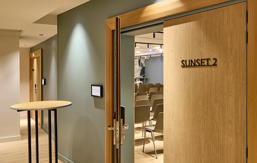 Entrée de la salle de réunion Sunset 2 de l'hôtel Le Talaia Mgallery Biarritz, vue depuis le couloir aux murs vert sauge, avec porte coulissante en bois clair entrouverte laissant apercevoir des rangées de chaises en configuration théâtre et une table haute en arrière-plan