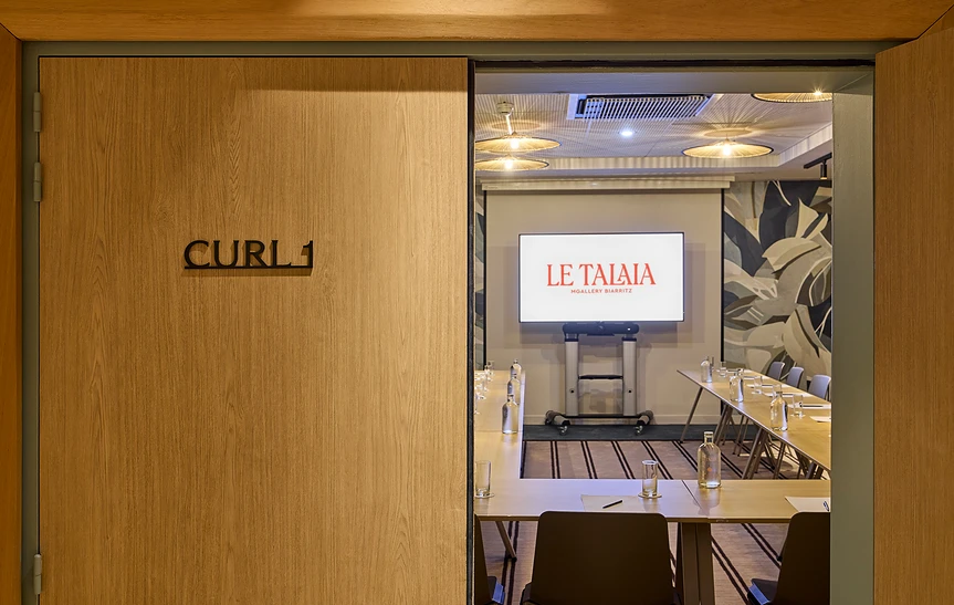 Entrée de la salle de réunion Curl de l'hôtel Le Talaia Mgallery Biarritz, vue depuis le couloir à travers une porte coulissante en bois clair, avec tables en U, verres d'eau et écran de projection au fond
