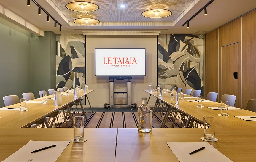 Salle de réunion de l'hôtel Le Talaia Mgallery Biarritz configurée en U, avec tables en bois clair dressées de carafes d'eau, verres et blocs-notes, écran de projection au centre, luminaires ronds en rotin et fresque murale végétale en arrière-plan
