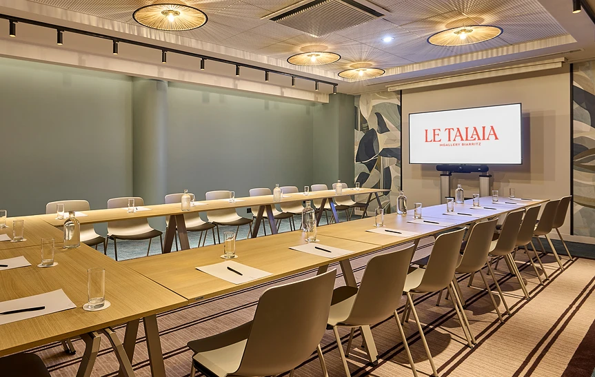 Salle de réunion de l'hôtel Le Talaia Mgallery Biarritz configurée en U, avec tables en bois clair et chaises grises, dressées de carafes d'eau, verres et feuilles, écran mural à droite, luminaires ronds en rotin au plafond et murs vert sauge