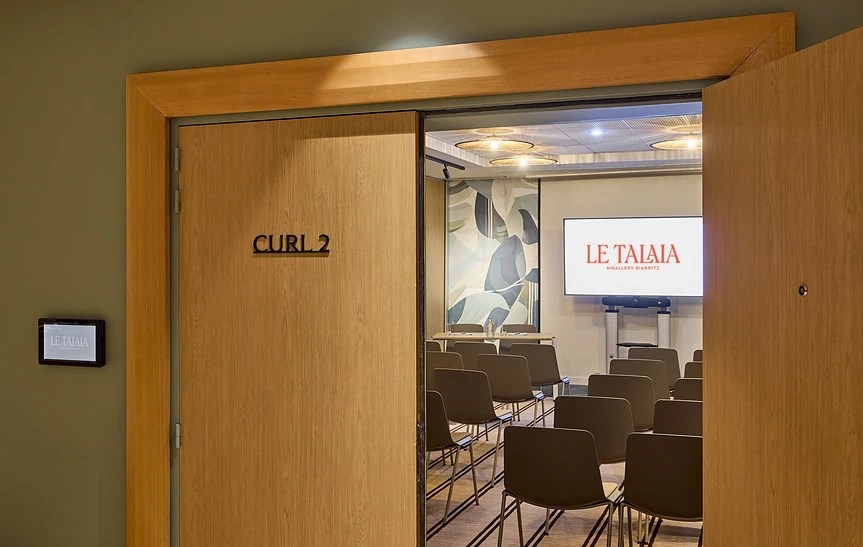 Entrée de la salle de réunion « Curl 2 » de l'hôtel Le Talaia MGallery Biarritz, avec porte coulissante en chêne ouverte sur une salle en configuration théâtre, écran de projection sur roulettes affichant le logo et fresque murale végétale en fond de salle.
