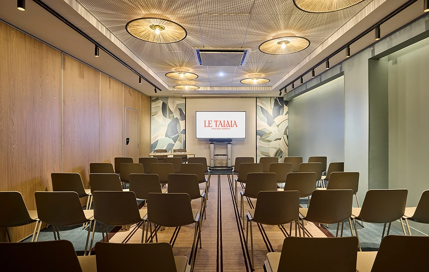 Salle de conférence de l'hôtel Le Talaia, avec rangées de chaises en théâtre face à un écran de projection, murs en bois clair, luminaires ronds en rotin au plafond et décoration murale végétale