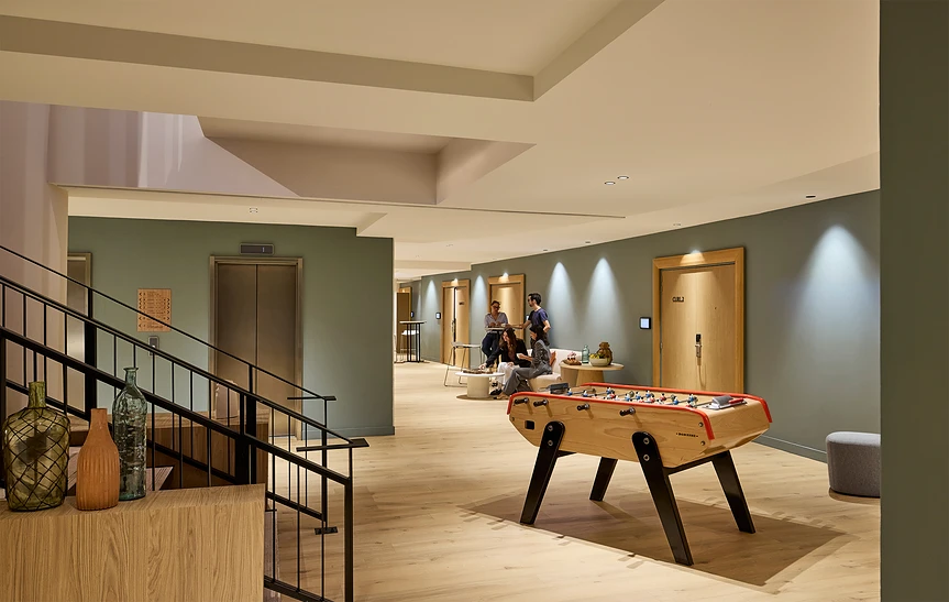Couloir de l'espace séminaire de l'hôtel Le Talaia MGallery Biarritz avec un baby-foot en bois, murs vert sauge, parquet clair, escalier métallique, vases décoratifs et groupe de participants en discussion près des salles de réunion.