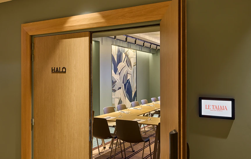Couloir de l'espace séminaire de l'hôtel Le Talaia MGallery Biarritz avec porte coulissante en chêne de la salle « Halo » entrouverte sur une table dressée et une fresque végétale, et écran digital affichant le logo de l'hôtel sur le mur vert kaki.