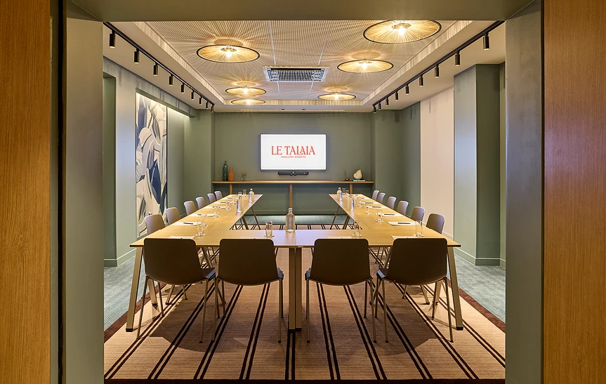 Salle de réunion « Halo » de l'hôtel Le Talaia MGallery Biarritz vue depuis l'entrée, en configuration en U avec tables dressées pour une vingtaine de personnes, murs vert sauge, plafond à suspensions en rotin, écran mural et fresque végétale abstraite.