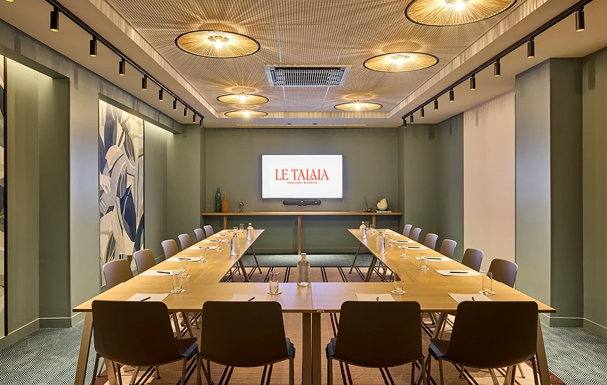 Vue intérieure de la salle « Halo » de l'hôtel Le Talaia MGallery Biarritz en configuration en U, avec tables dressées, murs vert kaki, nombreuses suspensions en rotin au plafond, étagère décorative avec vases et fresque murale végétale sur le côté.