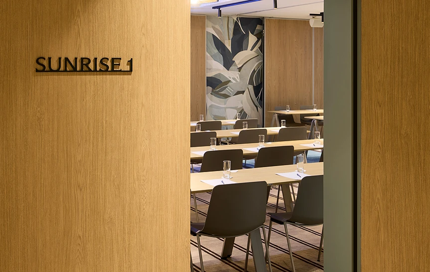 Entrée de la salle de séminaire « Sunrise 1 » de l'hôtel Le Talaia MGallery Biarritz, vue depuis l'encadrement de porte en chêne, avec tables dressées en configuration classe, chaises taupe et fresque murale végétale abstraite en fond de salle.