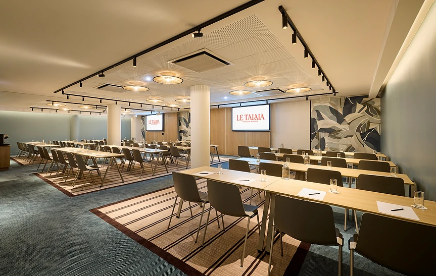 Vue d'ensemble de la grande salle de séminaire modulable de l'hôtel Le Talaia MGallery Biarritz ouverte sur deux espaces en configuration classe, avec deux écrans de projection, suspensions en rotin, cloisons acoustiques en chêne et fresque murale végétale en fond de salle.