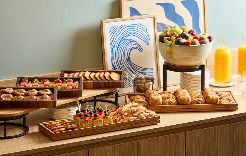 Buffet de pause séminaire soigneusement dressé sur un meuble en bois clair, avec plateaux de madeleines, fruits frais, viennoiseries, mini-sandwichs et brochettes salées, coupe de fruits frais, jus d'orange et illustration d'une vague en arrière-plan.