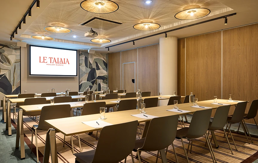 Salle de séminaire de l'hôtel Le Talaia MGallery Biarritz en configuration classe, avec rangées de tables en bois clair dressées, chaises taupe, plafond orné de suspensions en rotin, cloisons acoustiques en bois et écran de projection affichant le logo de l'hôtel.