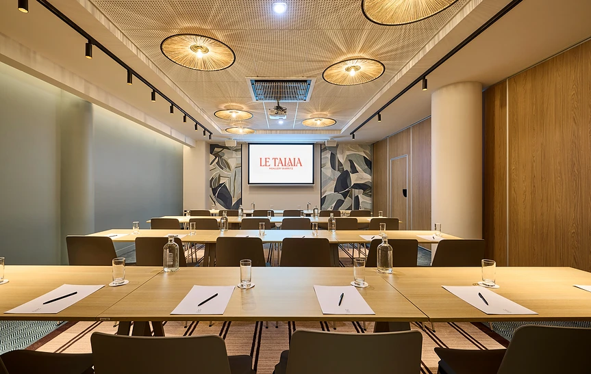 Salle de séminaire de l'hôtel Le Talaia MGallery Biarritz en configuration en U, avec tables dressées en bois clair, chaises taupe, plafond orné de suspensions en rotin, cloison acoustique en chêne, fresque murale végétale et écran de projection.
