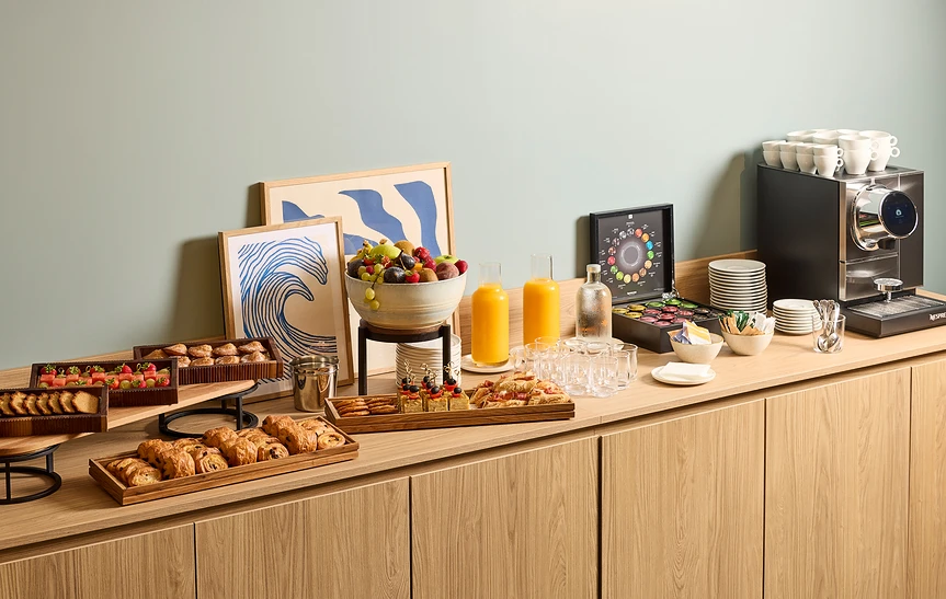 Buffet de pause café séminaire dressé sur un meuble en bois clair avec viennoiseries, fruits frais, mini-pâtisseries, jus d'orange, machine Nespresso, tasses et capsules, le tout mis en scène avec une illustration d'une vague bleue en arrière-plan.