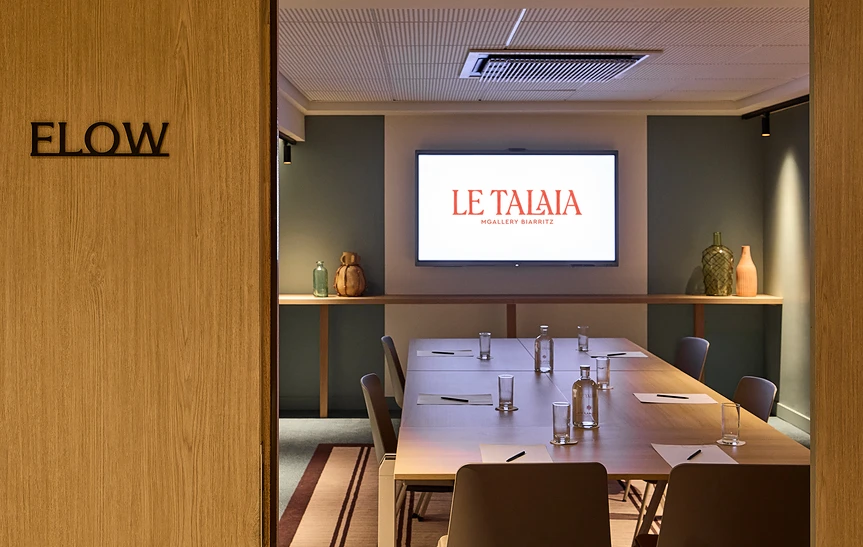 Salle de réunion « Flow » de l'hôtel Le Talaia MGallery Biarritz, vue depuis l'entrée en bois, avec table de réunion dressée pour une dizaine de personnes, écran mural affichant le logo de l'hôtel et étagère décorative avec vases en verre coloré.