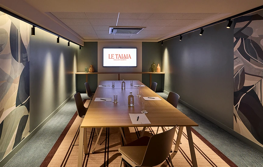 Salle de réunion feutrée de l'hôtel Le Talaia MGallery Biarritz avec table longue en bois clair, chaises en bois sombre, murs ornés de fresques végétales abstraites, éclairage sur rail et écran mural affichant le logo de l'hôtel.