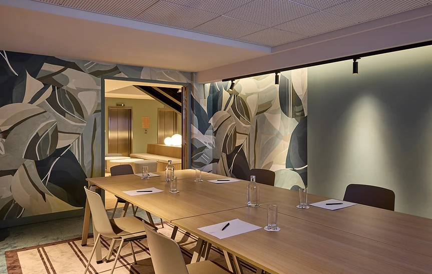 Salle de réunion modulable de l'hôtel Le Talaia MGallery Biarritz avec table en bois clair dressée, cloison mobile ouverte sur un espace adjacent, murs entièrement habillés d'un papier peint à motifs de feuilles tropicales en camaïeu bleu-vert et beige.