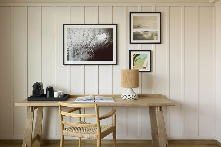 Bureau en chêne avec machine à café, livre ouvert et lampe, devant un mur lambrisé blanc orné de trois œuvres encadrées à thème marin.