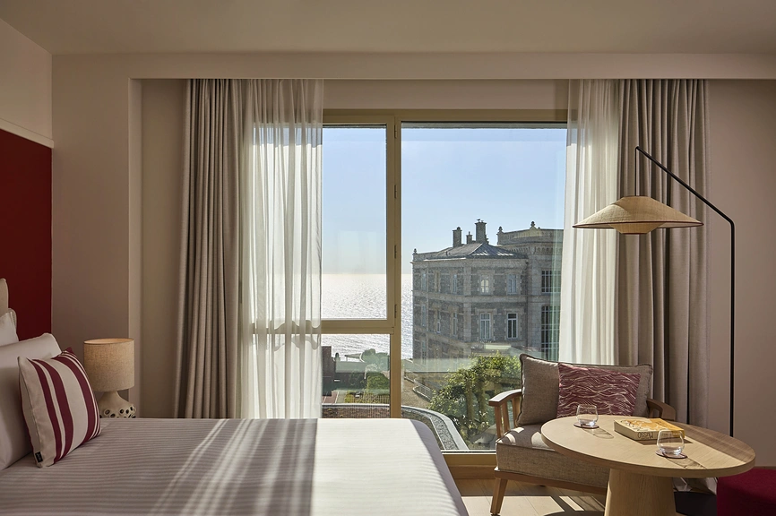 Chambre d'hôtel avec lit, fauteuil et grande baie vitrée offrant une vue sur un bâtiment historique en pierre et l'océan.