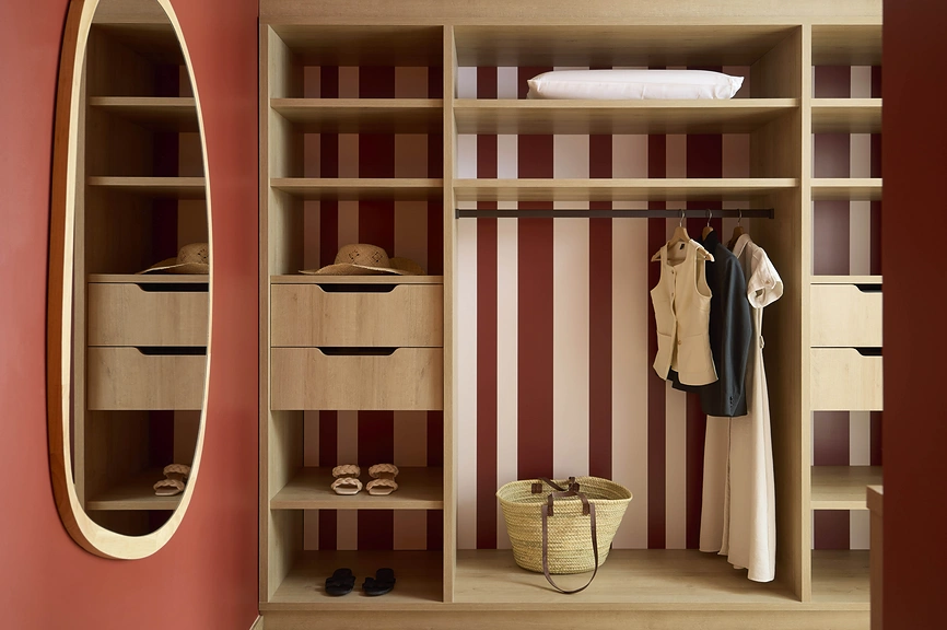 Dressing ouvert en chêne clair avec fond rayé bordeaux, tiroirs, penderie et étagères à chaussures, flanqué d'un miroir ovale sur mur terracotta.