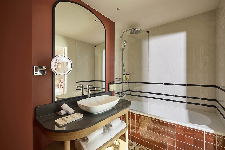 Salle de bain d'hôtel avec vasque à poser sur plan noir, baignoire-douche à carrelage terracotta, miroir arqué et miroir de courtoisie sur mur bordeaux.