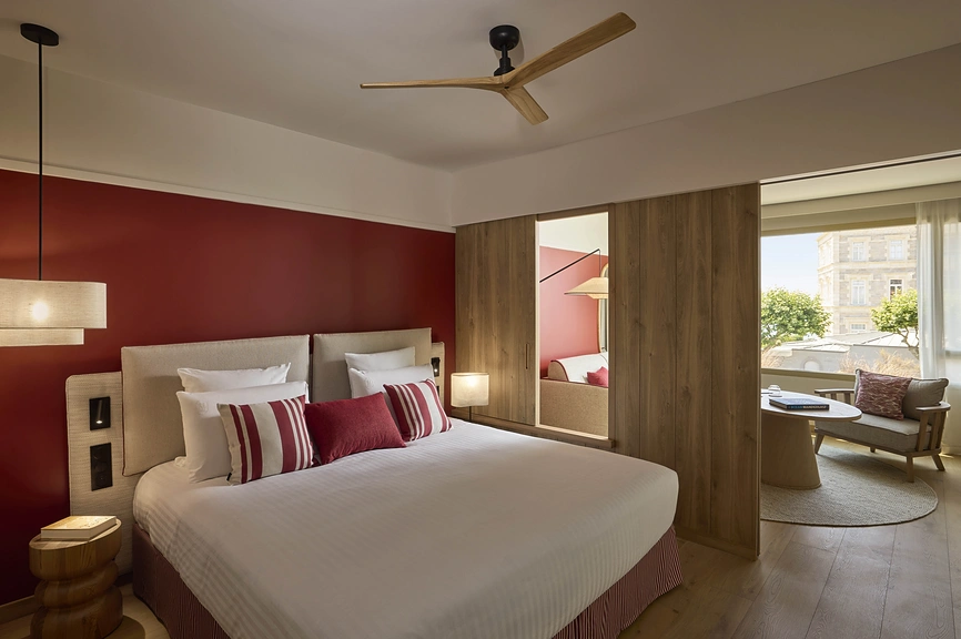 Chambre d'hôtel avec grand lit, coussins rayés bordeaux, mur rouge, cloison en bois ouvrant sur un salon lumineux avec vue sur la ville.